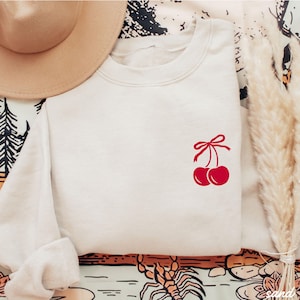 May include: A white sweatshirt with a red cherry graphic on the front. The cherries have a red bow on the stem.