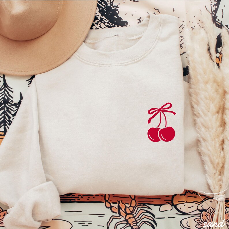 May include: A white sweatshirt with a red cherry graphic on the front. The cherries have a red bow on the stem.