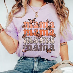 May include: A lavender t-shirt with a distressed graphic print of the word "mama" repeated four times in different colors and styles. The graphic is surrounded by orange butterflies.