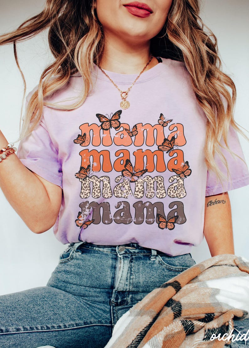 May include: A lavender t-shirt with a distressed graphic print of the word "mama" repeated four times in different colors and styles. The graphic is surrounded by orange butterflies.
