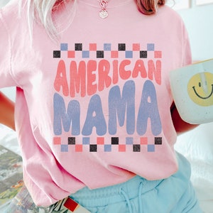May include: A pink t-shirt with the text "AMERICAN MAMA" in a retro font. The text is in shades of pink, blue, and black. The shirt has a checkered pattern around the text.