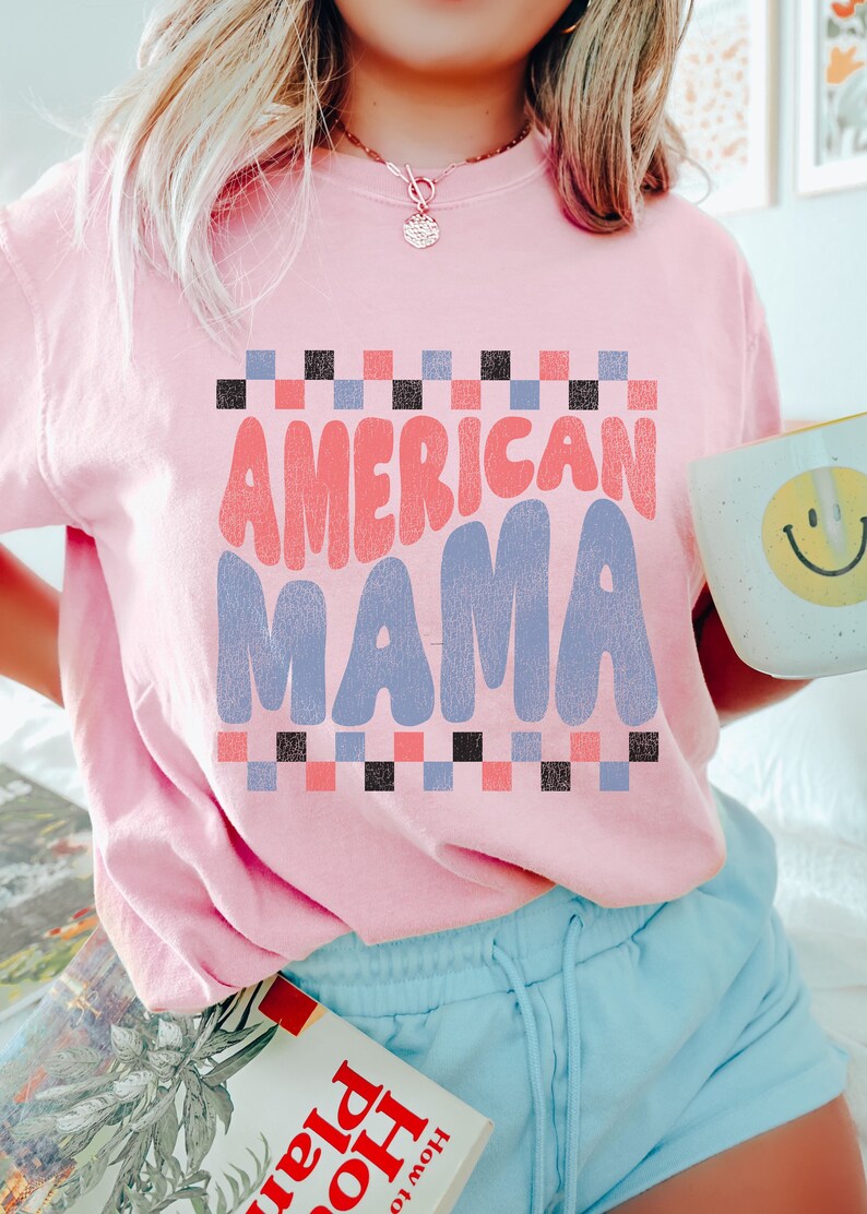 May include: A pink t-shirt with the text "AMERICAN MAMA" in a retro font. The text is in shades of pink, blue, and black. The shirt has a checkered pattern around the text.
