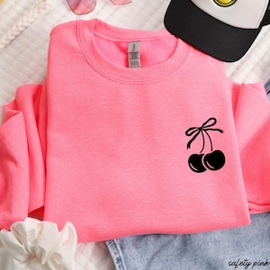May include: A pink sweatshirt with a black cherry design on the front. The cherries have a black bow on top.