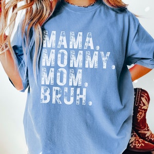 May include: A blue t-shirt with the text "MAMA. MOMMY. MOM. BRUH." printed in white on the front. The text has a distressed, vintage look.