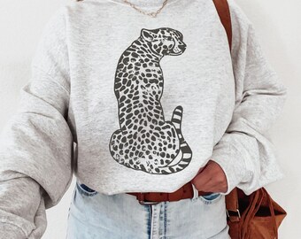 Può includere: Una felpa grigia con un design a stampa di ghepardo in bianco e nero. La felpa ha un collo rotondo e maniche lunghe.