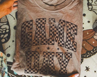 Pode incluir: Uma camiseta marrom com um design de padrão xadrez que diz "GAME DAY" em letras pretas.