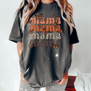 May include: A dark gray t-shirt with the text "mama mama mama mama" in a retro font with orange, brown, and white butterflies.
