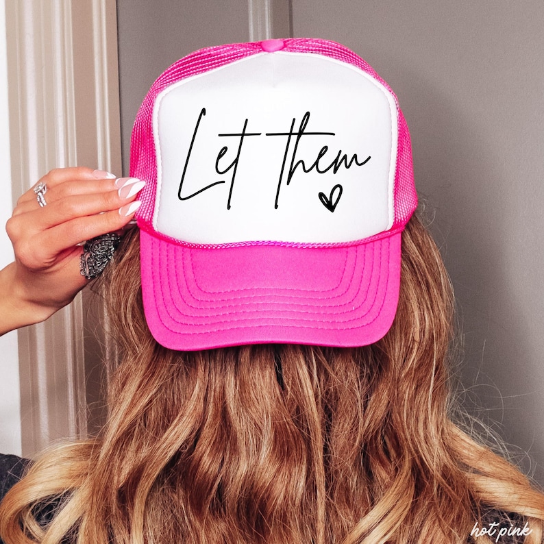 May include: A hot pink trucker hat with a white front panel. The text "Let them" is written in black script, with a small heart below. The hat has a mesh back and a curved brim.