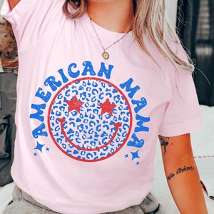 May include: Pink t-shirt with a blue and red smiley face graphic that says "American Mama" in a retro font. The smiley face has a leopard print pattern and stars around it.