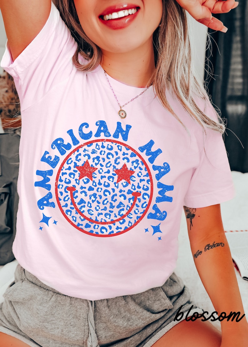 May include: Pink t-shirt with a blue and red smiley face graphic that says "American Mama" in a retro font. The smiley face has a leopard print pattern and stars around it.