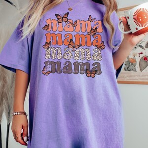 May include: Purple t-shirt with a distressed graphic print of the word "mama" repeated four times in a retro font. The graphic is surrounded by orange butterflies.