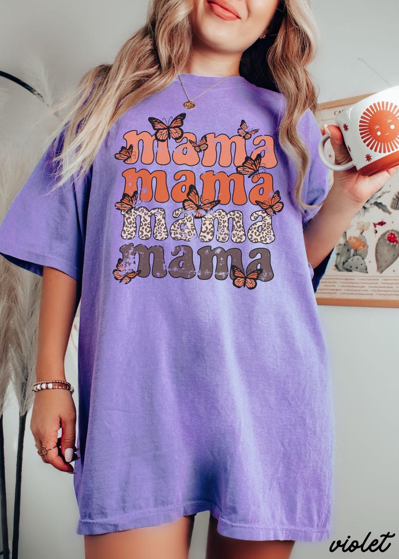 May include: Purple t-shirt with a distressed graphic print of the word "mama" repeated four times in a retro font. The graphic is surrounded by orange butterflies.