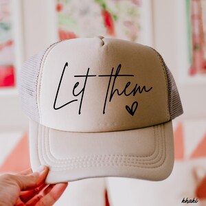 May include: Khaki trucker hat with the phrase "Let them" in black script and a small heart. The hat has a mesh back and a curved brim. The word "khaki" is printed in the bottom right corner.