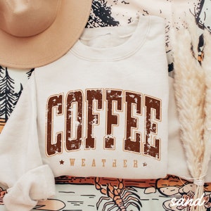 May include: A cream-colored sweatshirt with a brown distressed graphic that reads "COFFEE WEATHER".