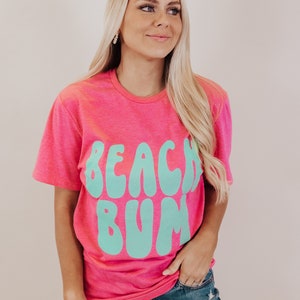 K&ouml;nnte beinhalten: Ein pinkfarbenes T-Shirt mit einer t&uuml;rkisblauen Grafik, auf der in einer Retro-Schrift "Beach Bum" steht.