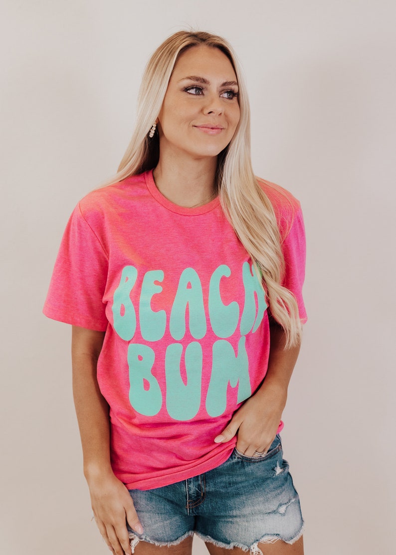 K&ouml;nnte beinhalten: Ein pinkfarbenes T-Shirt mit einer t&uuml;rkisblauen Grafik, auf der in einer Retro-Schrift "Beach Bum" steht.