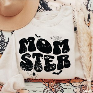 May include: A cream-colored sweatshirt with a black graphic that says "MOMSTER" in a retro font. The graphic also includes a spider, a spiderweb, and bats.