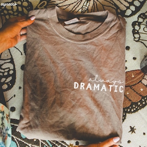 May include: A brown t-shirt with the text "always dramatic" printed in white on the front.