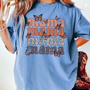 May include: A blue t-shirt with a graphic design that says "mama mama mama mama" in a retro font with butterflies and leopard print accents.