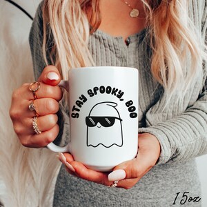 May include: White ceramic mug with the text "STAY SPOOKY, BOO" and a cartoon ghost wearing sunglasses. The mug is held by a person. The mug is 15 ounces.