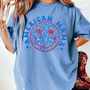 May include: A blue t-shirt with a red, white, and blue smiley face graphic. The smiley face has stars for eyes and a leopard print pattern. The text "American Mama" is written around the smiley face.