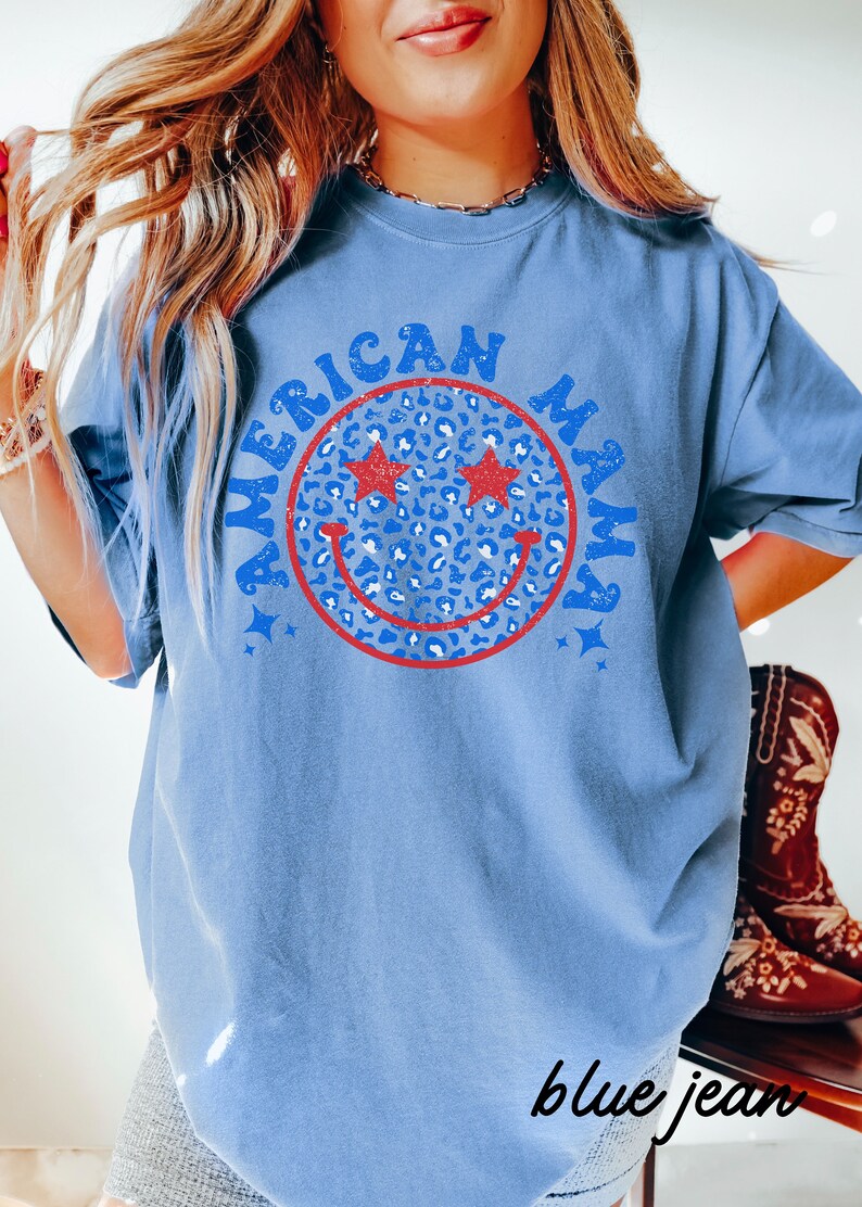 May include: A blue t-shirt with a red, white, and blue smiley face graphic. The smiley face has stars for eyes and a leopard print pattern. The text "American Mama" is written around the smiley face.