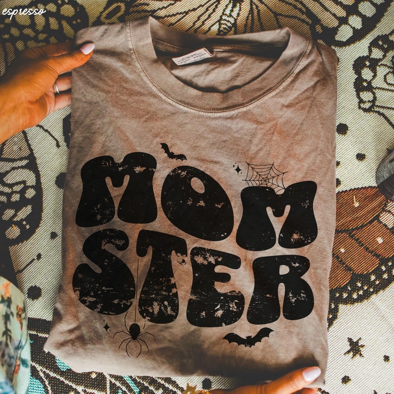 May include: A brown t-shirt with a distressed black graphic that says "MOMSTER" in a retro font. The graphic also includes a spider, a spider web, and bats.