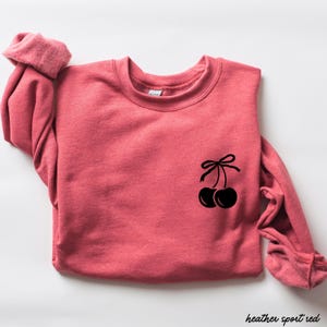 May include: A heather sport red sweatshirt with a black cherry graphic on the front. The cherries have a stem with a bow on top.