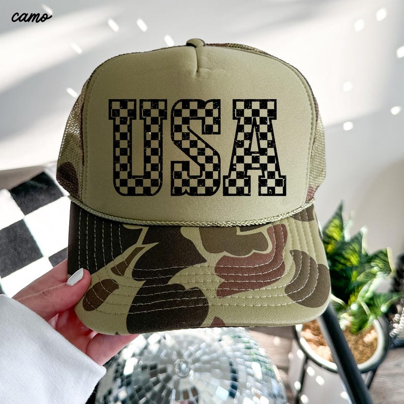 May include: A beige trucker hat with a camouflage pattern on the brim. The front of the hat features a black and white checkered design that spells out "USA".