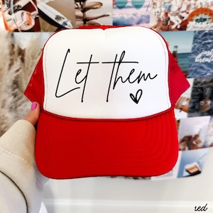 May include: Red and white trucker hat with the text "Let them" in black script and a small heart. The hat has a red brim and mesh sides, with a white front panel.
