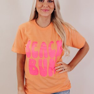 K&ouml;nnte beinhalten: Ein pfirsichfarbenes T-Shirt mit dem Schriftzug "Beach Bum" in pink.
