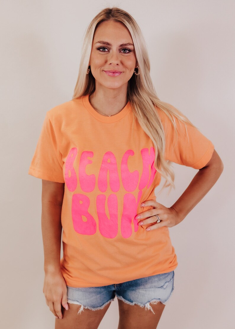 K&ouml;nnte beinhalten: Ein pfirsichfarbenes T-Shirt mit dem Schriftzug "Beach Bum" in pink.