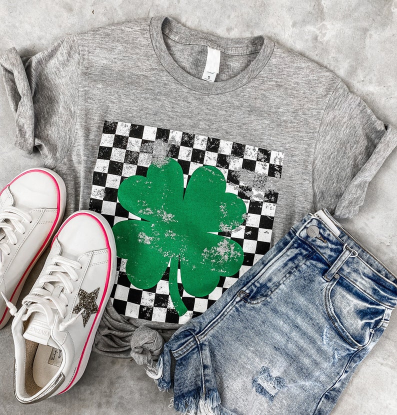 May include: A gray t-shirt with a green four-leaf clover graphic on a black and white checkered background. The clover has a distressed look. The shirt is paired with a light blue denim shorts.