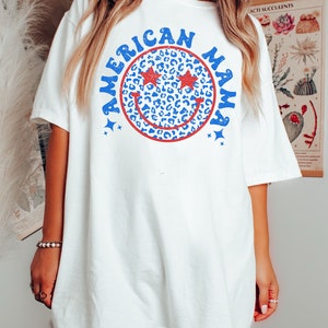 May include: White oversized t-shirt with a blue and red smiley face graphic that says "American Mama". The smiley face has red stars for eyes and a leopard print pattern.