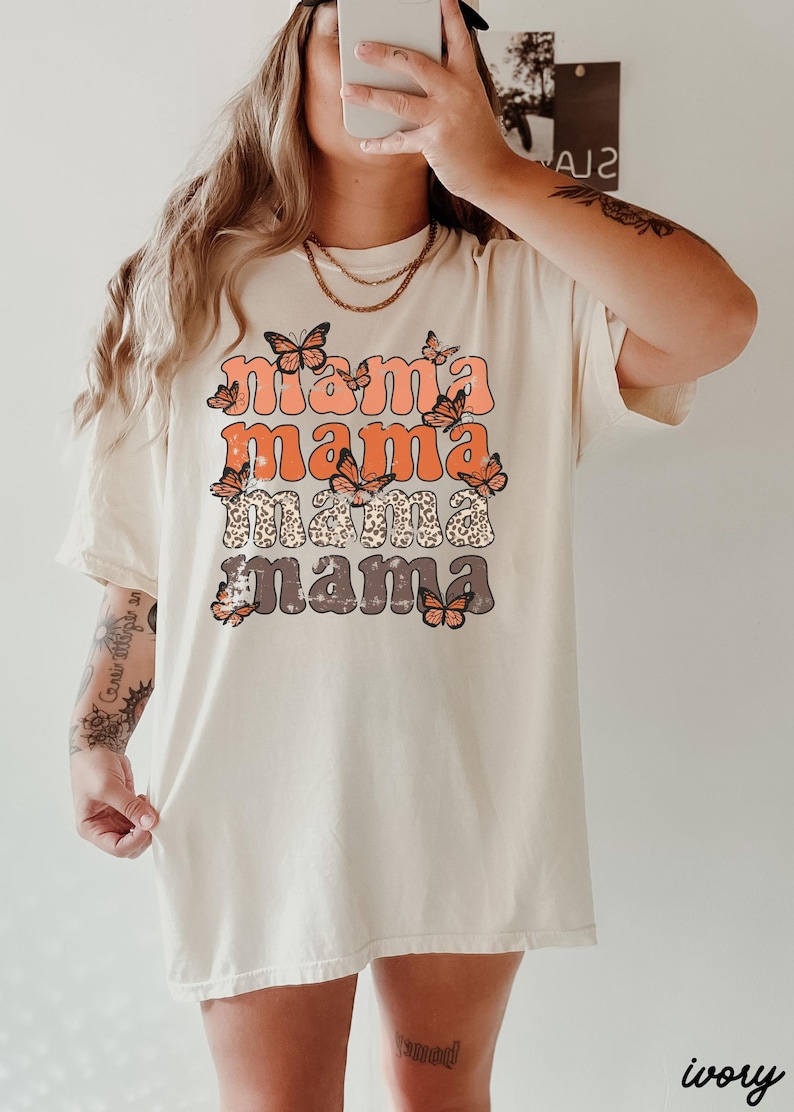 May include: A white t-shirt with a graphic print of the word "mama" repeated four times in a stacked design. Each word is in a different color and has a butterfly design around it. The shirt is a relaxed fit and has a crew neck.