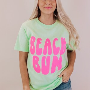 K&ouml;nnte beinhalten: Ein hellgr&uuml;nes T-Shirt mit einem pinkfarbenen Aufdruck, der in einer Retro-Schrift "Beach Bum" sagt.
