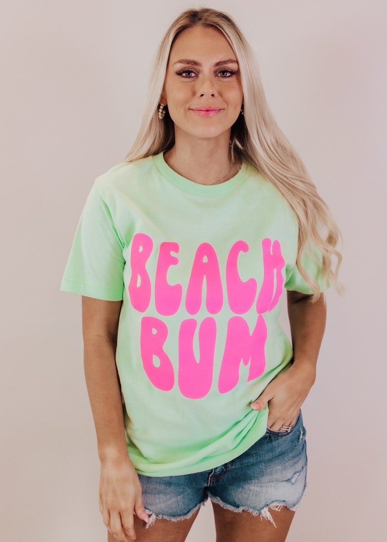 K&ouml;nnte beinhalten: Ein hellgr&uuml;nes T-Shirt mit einem pinkfarbenen Aufdruck, der in einer Retro-Schrift "Beach Bum" sagt.