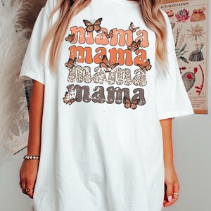 May include: White oversized t-shirt with a graphic design of the word "mama" repeated four times in different colors and styles. The word "mama" is surrounded by butterfly graphics.