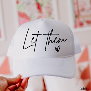 May include: White trucker hat with the text "Let them" in a black script font, accompanied by a small heart symbol. The hat features a mesh back and a curved brim. The hat is held by a person's hand.
