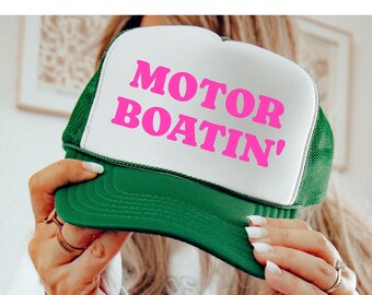 Può includere: Un cappellino da camionista verde e bianco con la scritta "MOTOR BOATIN'" in lettere rosa acceso. Il cappello ha un retro in rete e una visiera curva. Un accessorio divertente.