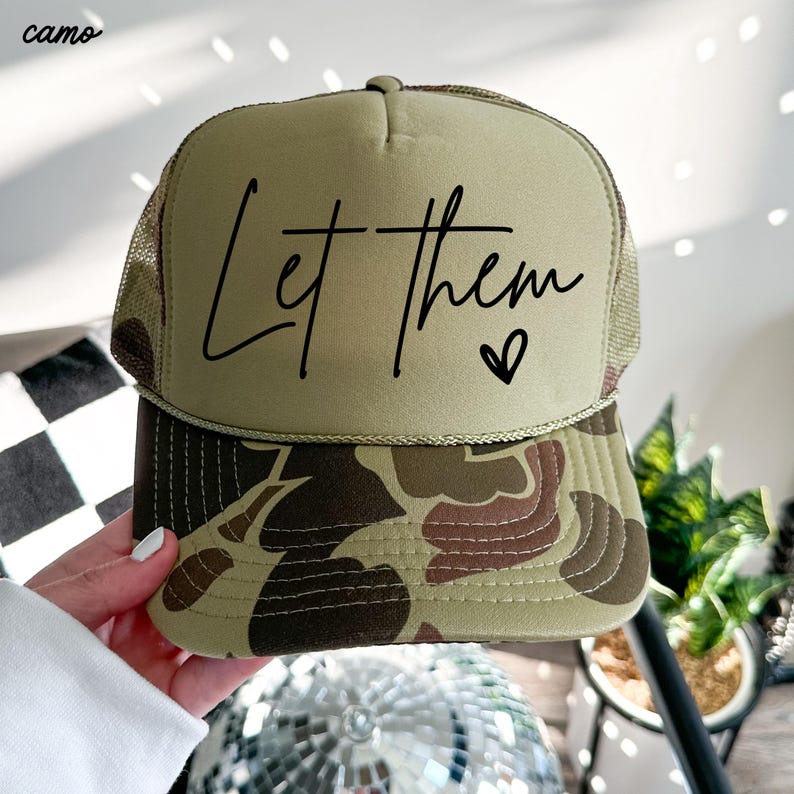 May include: A trucker hat with a camo brim and mesh back. The front panel is tan with the words "Let them" and a heart symbol in black script. The hat is a neutral color.