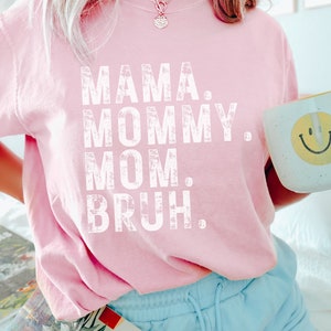 May include: A pink t-shirt with the text "MAMA. MOMMY. MOM. BRUH." printed in white. The shirt is worn by a person with long blonde hair and is paired with light blue shorts.