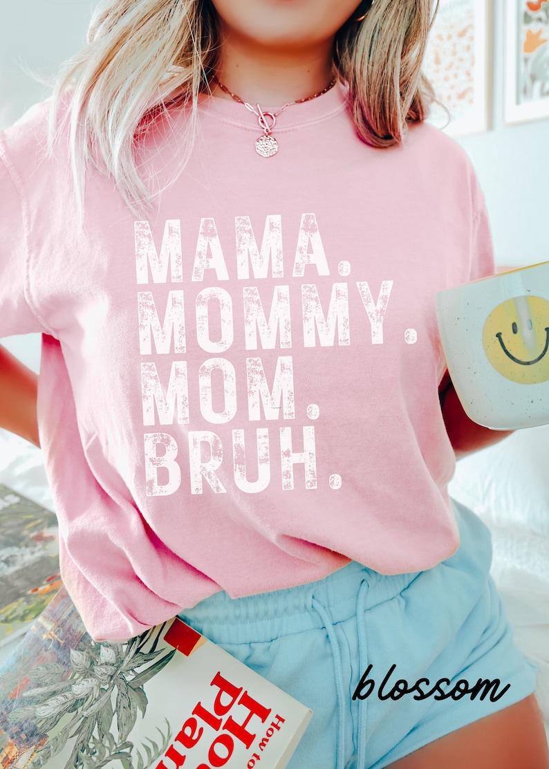 May include: A pink t-shirt with the text "MAMA. MOMMY. MOM. BRUH." printed in white. The shirt is worn by a person with long blonde hair and is paired with light blue shorts.