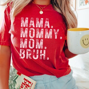 May include: Red t-shirt with the text "Mama. Mommy. Mom. Bruh." in pink lettering. The shirt is worn by a person wearing blue shorts with the word "red" written on the side.