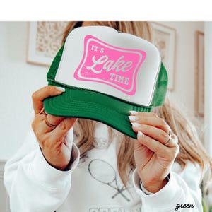 May include: A green and white trucker hat with the text "IT'S Lake TIME" in pink. The hat has a green brim and mesh back, and a white front panel. The text is inside a pink rectangle.