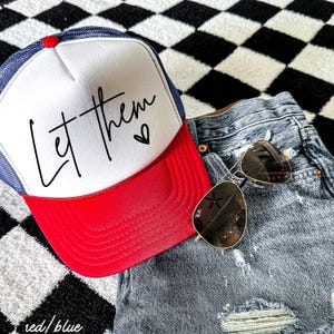 May include: A red, white, and blue trucker hat with the text "Let Them" in black script and a small heart. The hat is paired with denim shorts and sunglasses.