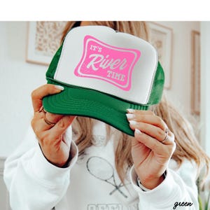 May include: Green and white trucker hat with the text "IT'S River TIME" in pink. The hat has a green brim and mesh back, and a white front panel with the pink text.