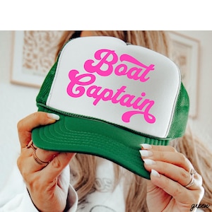 May include: A green and white trucker hat with the words "Boat Captain" in bright pink script across the front. The hat has a mesh back and a curved brim. The hat is a fun accessory.