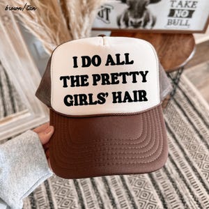May include: A brown and tan trucker hat with the text "I DO ALL THE PRETTY GIRLS' HAIR" in black. The hat has a brown brim and a white front panel. The mesh backing is tan.