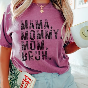 May include: A woman wearing a purple t-shirt with the text "MAMA. MOMMY. MOM. BRUH." in black distressed lettering. She is holding a mug and a book titled "House Plants".